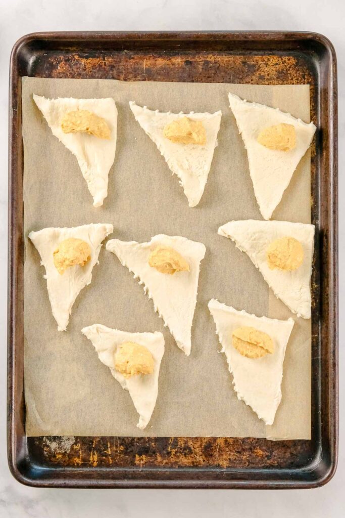 Triangles of crescent dough with a dollop of pumpkin crease cheese filling on each are arranged on a parchment-lined baking sheet.