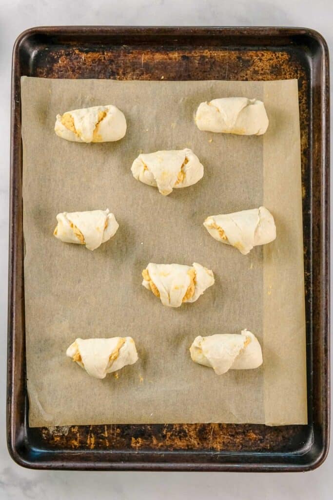 Eight unbaked crescent roll pastries with filling are arranged on a parchment-lined baking sheet.