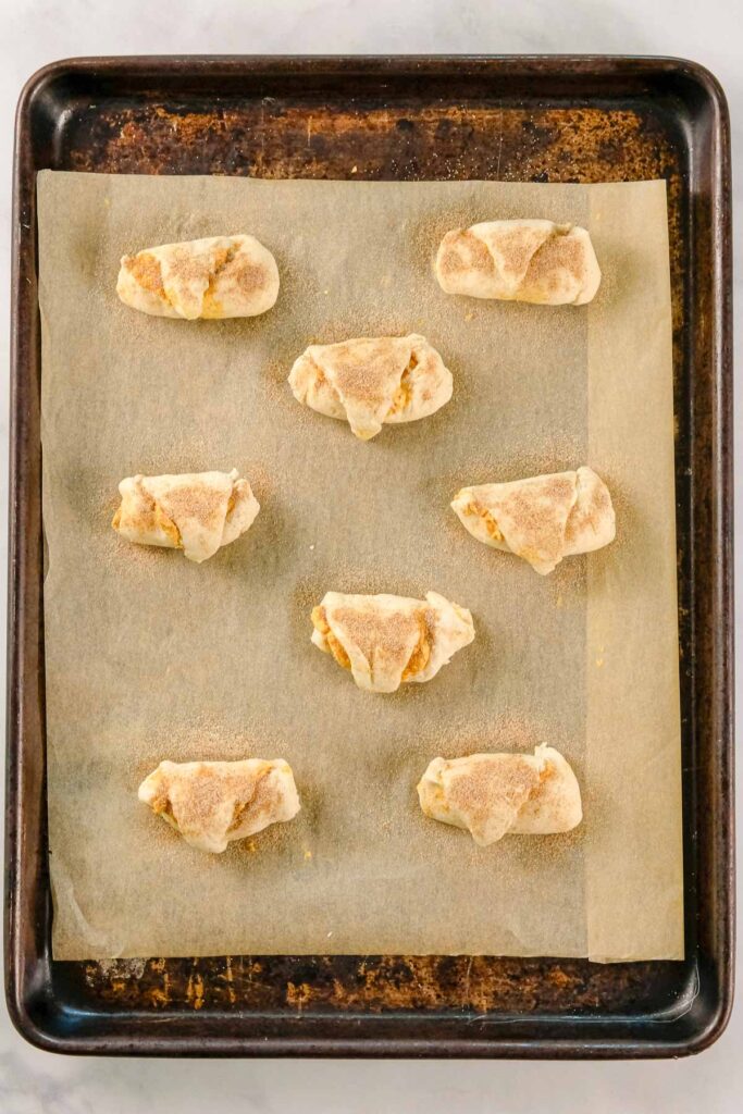 Eight unbaked crescent-shaped pastries topped with brown sugar are arranged on a parchment-lined baking sheet, ready to be baked.