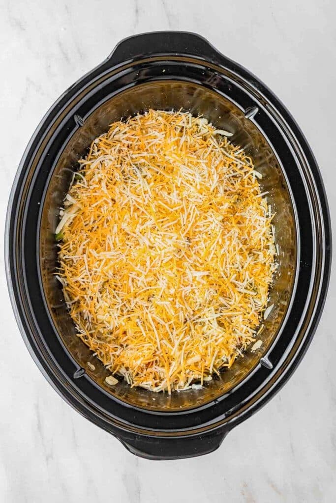 A slow cooker filled with shredded cheese on top of a casserole mixture, viewed from above on a marble countertop.