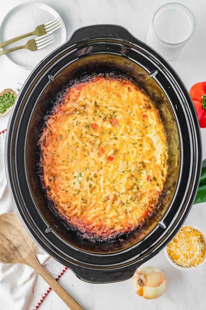 A slow cooker breakfast casserole topped with herbs sits in a black slow cooker, surrounded by plates, a glass, utensils, and vegetables on a white surface.