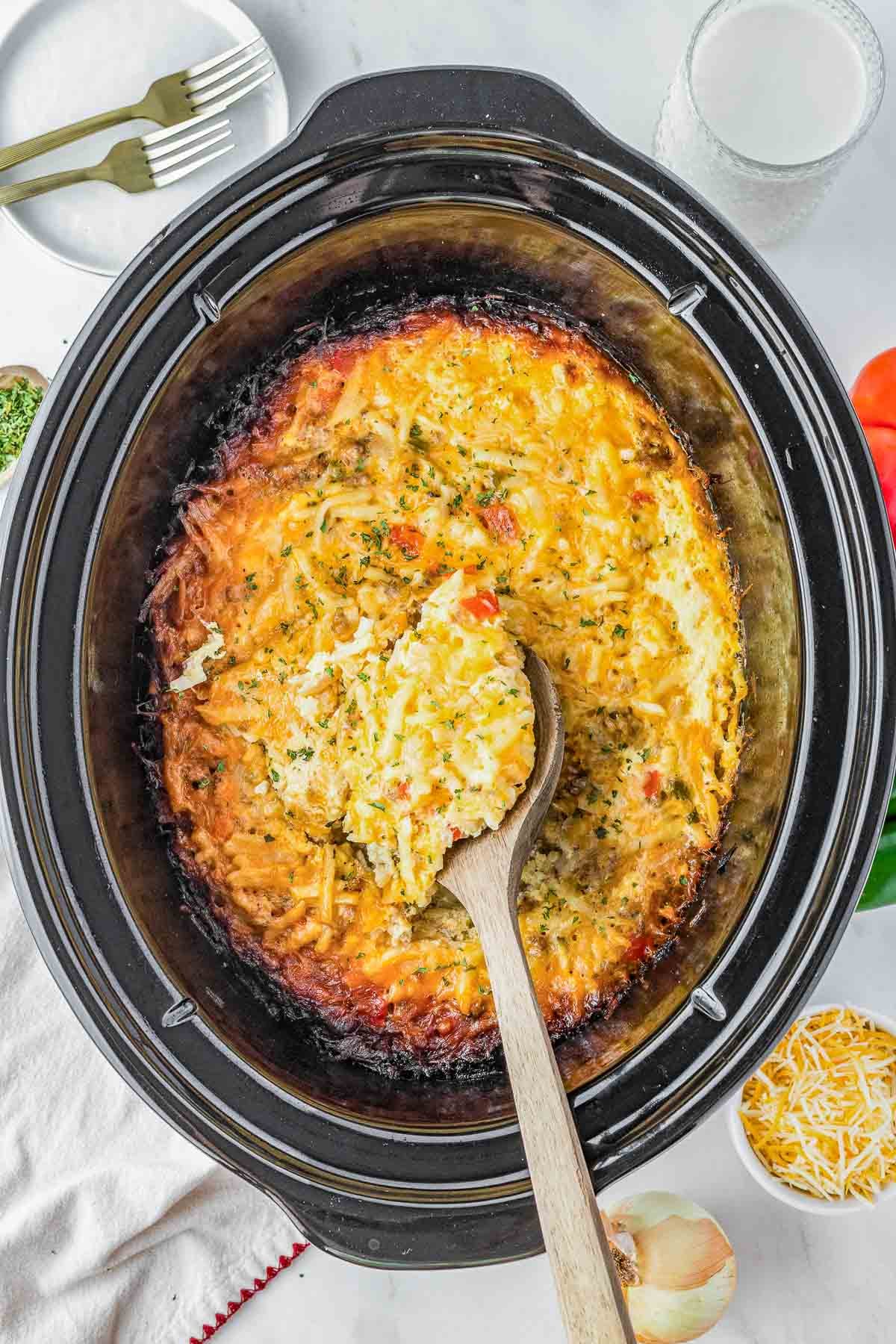 A wooden spoon scoops slow cooker breakfast casserole from a slow cooker; shredded cheese, onion, and bell pepper are nearby.