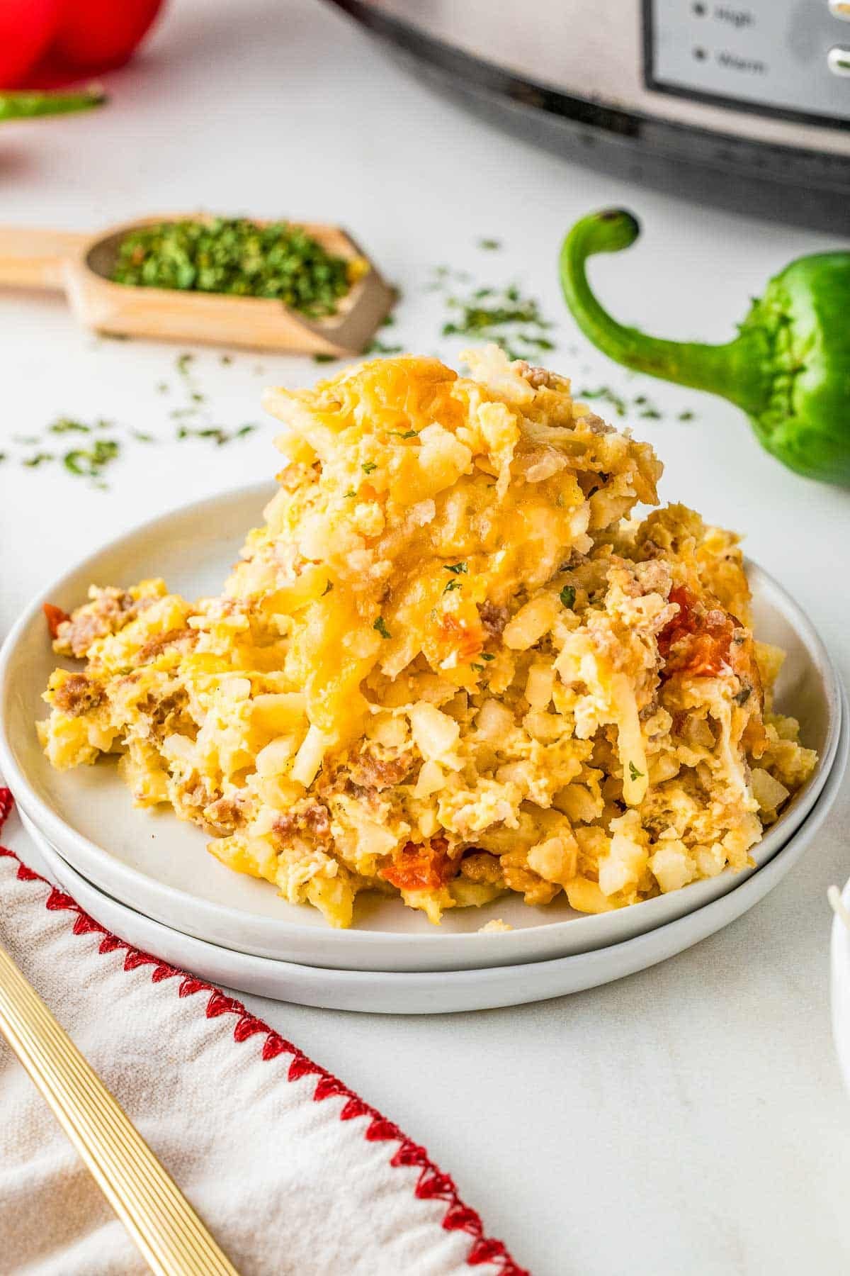 A serving of cheesy slow cooker breakfast casserole with ground meat and vegetables is plated on a white dish, with a slow cooker and green bell pepper in the background.