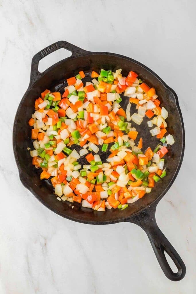 Diced onions, green bell peppers, and red bell peppers being sautéed in a black cast iron skillet on a light surface.