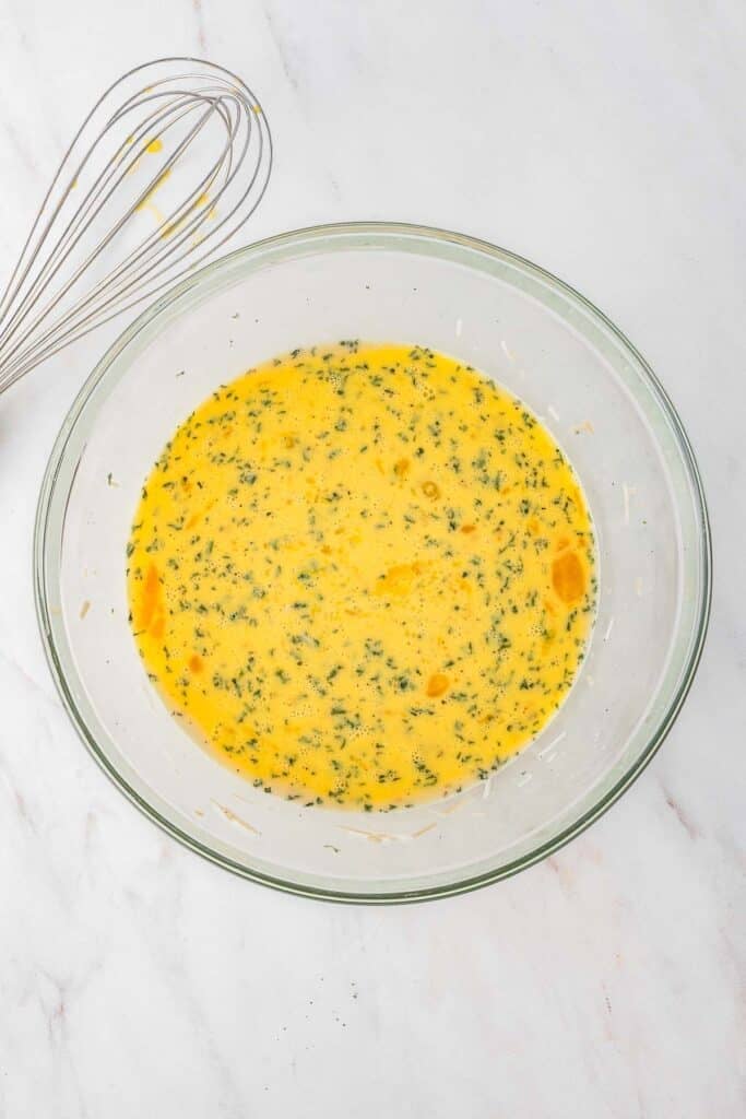 A glass bowl filled with a whisked egg and herb mixture sits on a white surface next to a metal whisk.