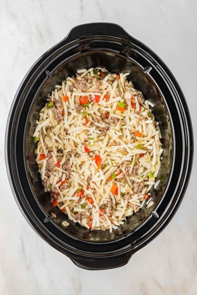 A slow cooker filled with a mixture of shredded potatoes, ground meat, diced red and green bell peppers, and onions, ready to be cooked.