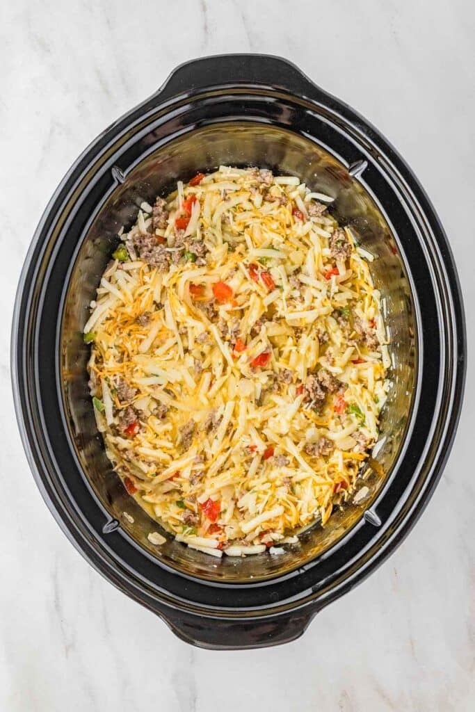 Slow cooker filled with a mixture of shredded cheese, hash browns, ground meat, and chopped vegetables on a white marble surface.