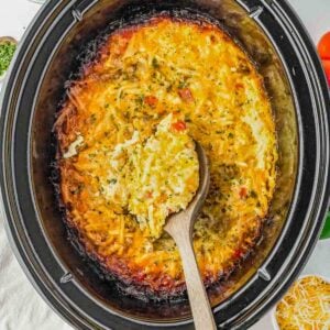 A wooden spoon scoops cheesy hash brown casserole from a black slow cooker; shredded cheese garnish is visible nearby.