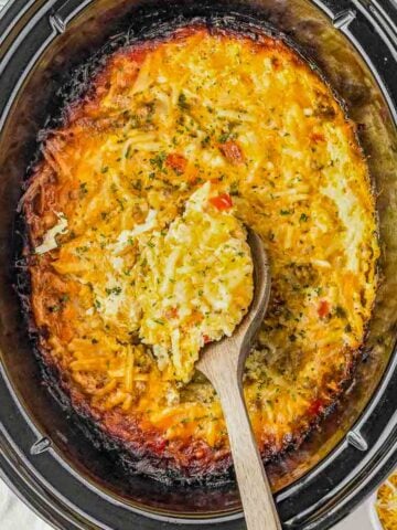 A wooden spoon scoops cheesy hash brown casserole from a black slow cooker; shredded cheese garnish is visible nearby.